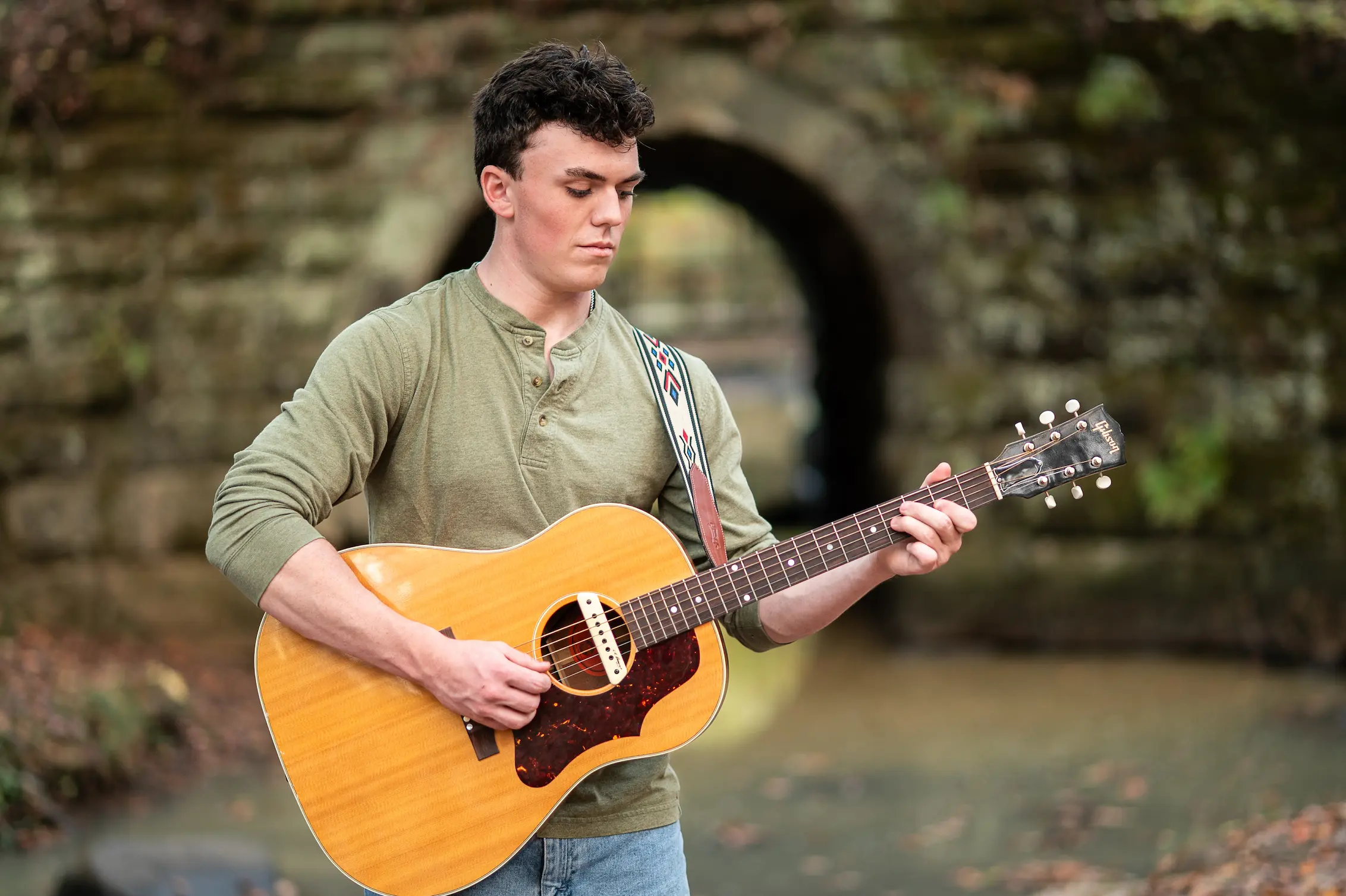 A senior portrait of a young musician with an acoustic guitar beside a stone bridge